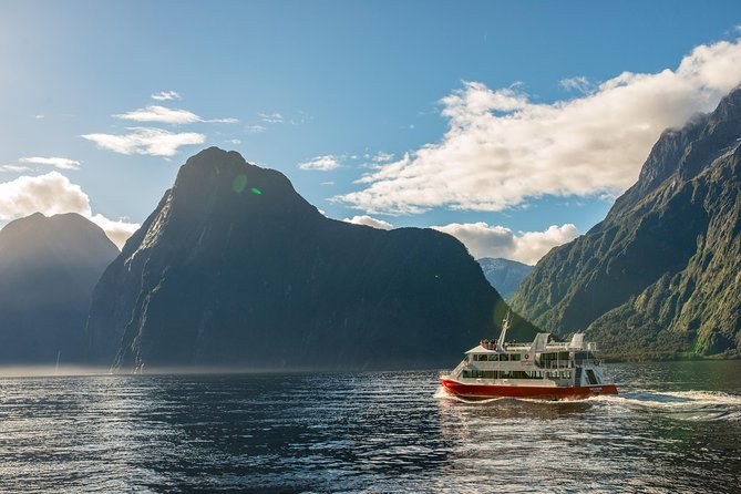 A red and white catamaran on dark water below bush-covered ridges and cloud