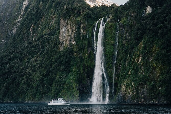 A small cruise boat below a long waterfall on a sheer green wall