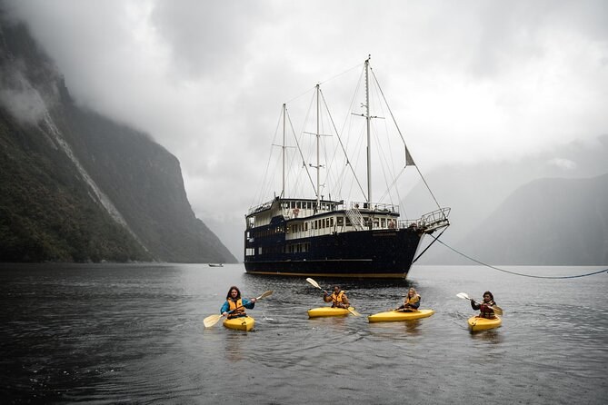 A tall ship at anchor on grey water, kayakers paddling in front of it