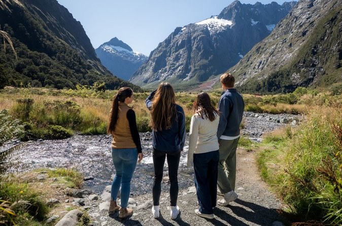 Four walkers on a riverside path in a glacier-cut valley under snowy peaks